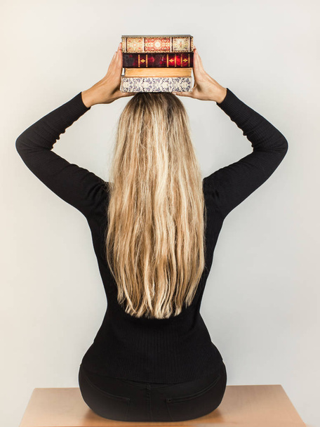 A blonde with long hair holds a stack of books on her head, sitt - Фото, изображение