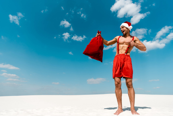 handsome and sexy man in santa hat pointing with finger at santa sack on beach in Maldives  - Foto, afbeelding