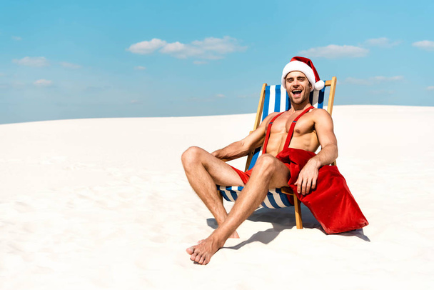 вродливий і сексуальний чоловік у капелюсі в Санті з мішком і сидячи на палубному стільці на пляжі Мальдівів.  - Фото, зображення