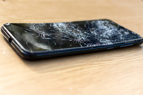 Broken screen cell phone on light  background. Old broken black smartphone on the table. Close up photos showing process of mobile phone repair. Smartphone and wooden hummer.  - 写真・画像
