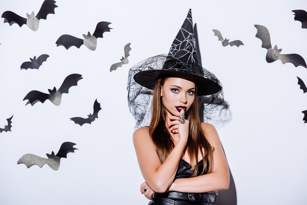 meisje in zwart heks Halloween kostuum kijken naar camera in de buurt van witte muur met decoratieve vleermuizen - Foto, afbeelding