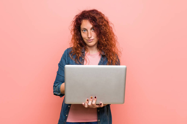 Fiatal kaukázusi vöröshajú nő, aki egy laptopot tart boldog, mosolygós és vidám. - Fotó, kép