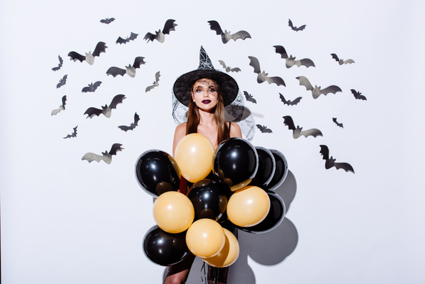 girl in black witch Halloween costume holding balloons near white wall with decorative bats - Photo, image