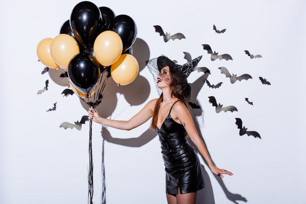happy girl in black witch Halloween costume holding balloons near white wall with decorative bats - Foto, Imagem