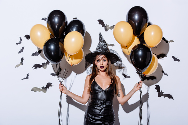 girl in black witch Halloween costume holding balloons near white wall with decorative bats - Photo, Image