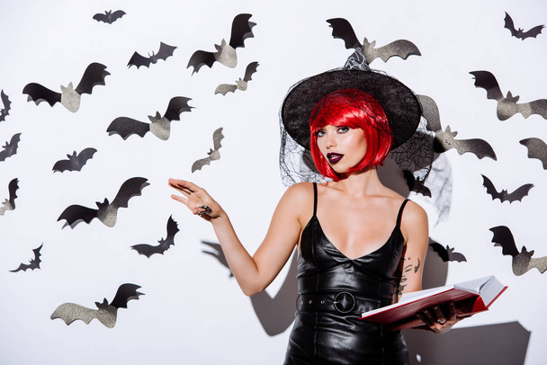 girl in black witch Halloween costume with red hair holding book near white wall with decorative bats - Foto, Bild