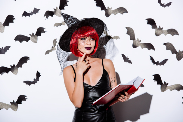 στοχαστικό κορίτσι σε μαύρο κοστούμι μάγισσα Απόκριες με κόκκινα μαλλιά κρατώντας βιβλίο κοντά σε λευκό τοίχο με διακοσμητικές νυχτερίδες - Φωτογραφία, εικόνα