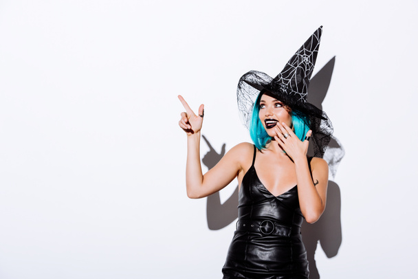smiling girl in black witch Halloween costume with blue hair pointing with finger on white background - Фото, изображение
