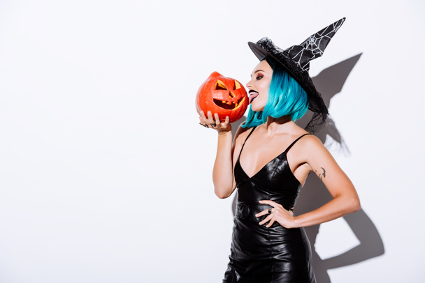 sexy girl in black witch Halloween costume with blue hair licking spooky carved pumpkin on white background - Fotografie, Obrázek