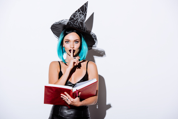 Siyah cadı cadılar bayramı kostümlü seksi kız elinde mavi saçla elinde kitap beyaz arka planda "şşş" hareketi gösteriyor. - Fotoğraf, Görsel