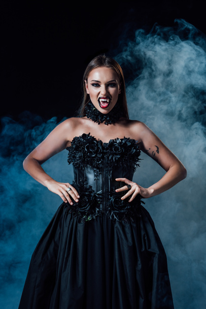 scary vampire girl with fangs in black gothic dress on black background with smoke - Zdjęcie, obraz