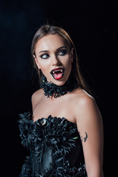 scary vampire girl with fangs in black gothic dress with open mouth looking away isolated on black - Foto, imagen
