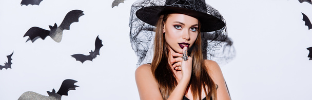 panoramisch schot van meisje in zwarte heks Halloween kostuum in de buurt van witte muur met decoratieve vleermuizen - Foto, afbeelding
