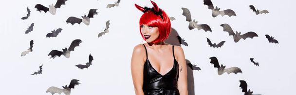 panoramisch schot van meisje in duivel Halloween kostuum in de buurt van witte muur met decoratieve vleermuizen - Foto, afbeelding