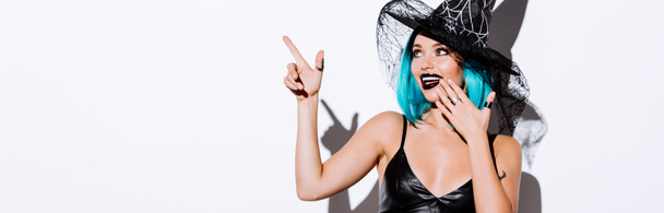 plano panorámico de chica sonriente en traje de Halloween bruja negro con el pelo azul señalando con el dedo sobre fondo blanco
 - Foto, Imagen
