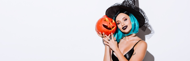 tiro panorâmico de menina sorridente em preto bruxa traje de Halloween com cabelo azul segurando assustador esculpida abóbora no fundo branco
 - Foto, Imagem