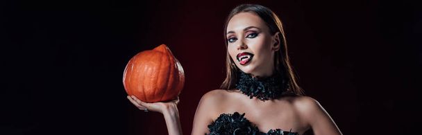 panoramisch schot van eng vampier meisje met hoektanden in zwart gotische jurk met Halloween pompoen op zwarte achtergrond - Foto, afbeelding