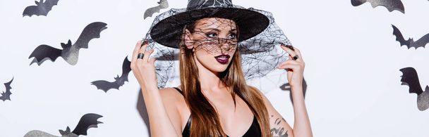 panoramic shot of girl in black witch Halloween costume holding veil on hat near white wall with decorative bats - Foto, Imagem
