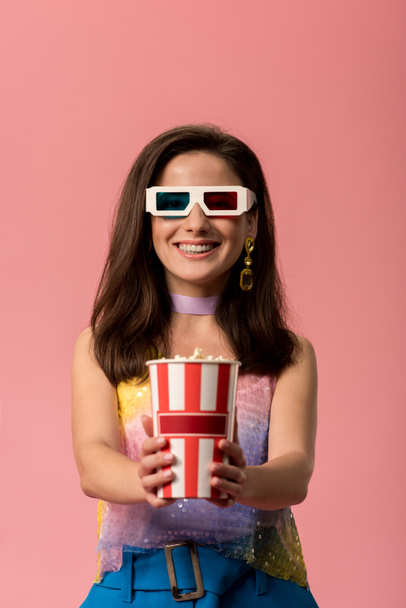 happy young stylish disco girl in 3d glasses holding striped paper bucket with popcorn isolated on pink - Foto, imagen