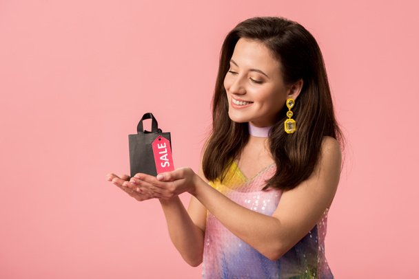 усміхнена стильна дівчина дискотека тримає сумку з продажем на етикетці ізольована на рожевому
 - Фото, зображення