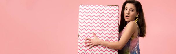 panoramic shot of shocked young stylish disco girl holding huge gift box isolated on pink - Photo, Image