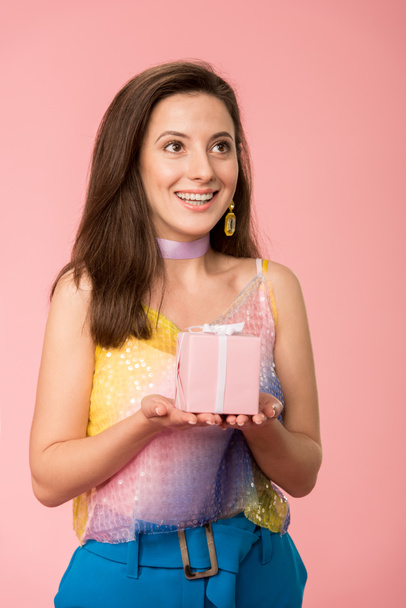 happy young stylish disco girl holding gift box and looking away isolated on pink - Foto, imagen