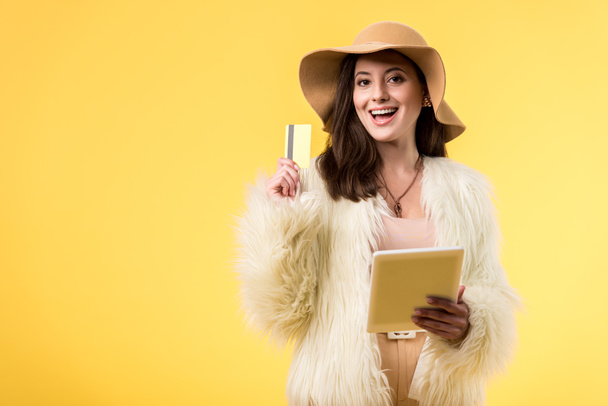 gelukkig elegant meisje in nep bont jas en hoed met digitale tablet en creditcard geïsoleerd op geel - Foto, afbeelding