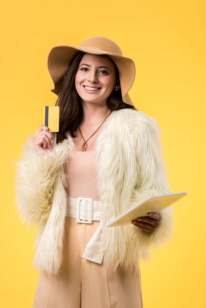 happy elegant girl in faux fur jacket and hat holding digital tablet and credit card isolated on yellow - Foto, immagini