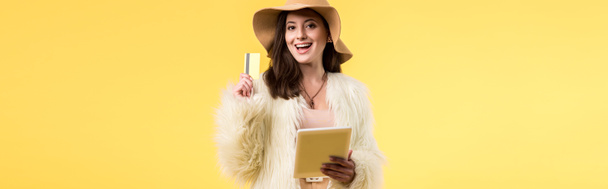 panoramic shot of happy elegant girl in faux fur jacket and hat holding digital tablet and credit card isolated on yellow - Fotó, kép