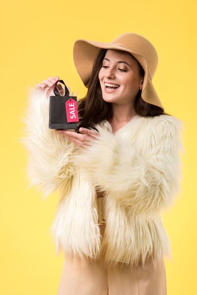 happy elegant girl in faux fur jacket and hat holding shopping bag with sale lettering isolated on yellow - Фото, изображение
