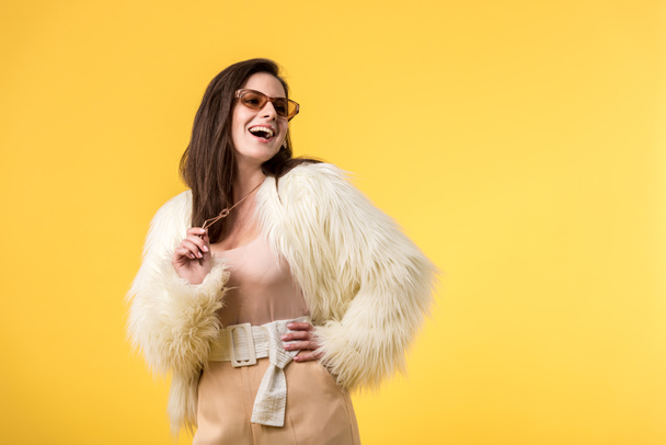 excited party girl in faux fur jacket and sunglasses isolated on yellow - Valokuva, kuva