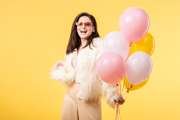 happy party girl in faux fur jacket and sunglasses holding balloons isolated on yellow - Foto, Bild