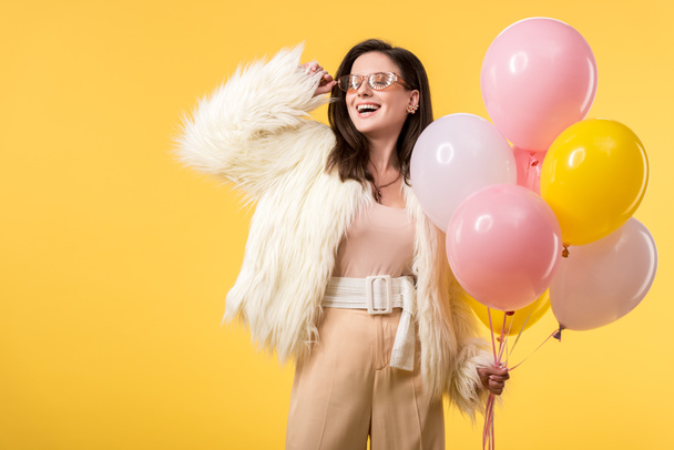 happy party girl in faux fur jacket and sunglasses holding balloons isolated on yellow - Zdjęcie, obraz