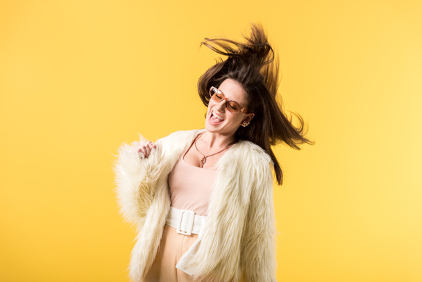 happy girl in faux fur jacket and sunglasses shaking head isolated on yellow - Photo, Image