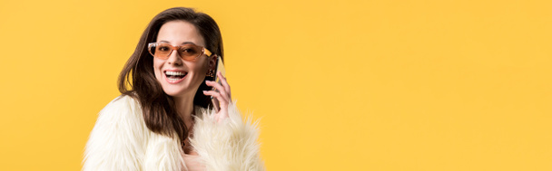 happy party girl in faux fur jacket and sunglasses talking on smartphone isolated on yellow, panoramic shot - Foto, immagini