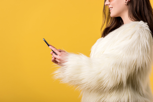 cropped view of happy party girl in faux fur jacket and sunglasses holding smartphone isolated on yellow - 写真・画像