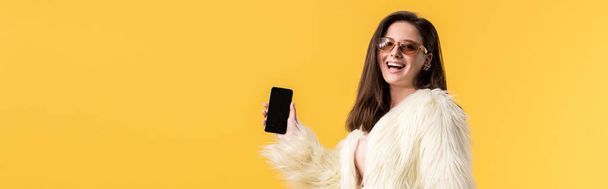 plano panorámico de chica de fiesta feliz en chaqueta de piel sintética y gafas de sol celebración de teléfono inteligente con pantalla en blanco aislado en amarillo
 - Foto, Imagen