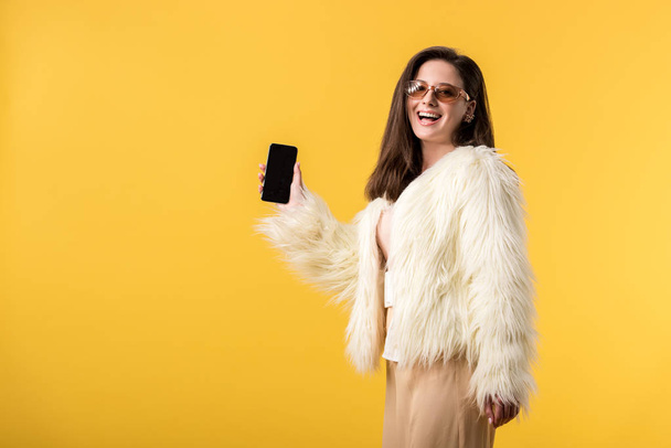 happy party dívka ve falešné kožešinové bundě a slunečních brýlích drží smartphone s prázdnou obrazovkou izolované na žluté - Fotografie, Obrázek