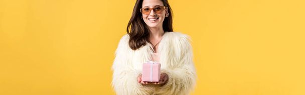 panoramic shot of happy party girl in faux fur jacket and sunglasses holding gift box isolated on yellow - Photo, Image