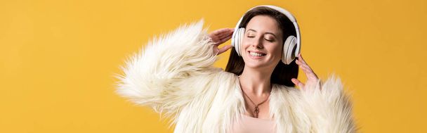 panoramiczne ujęcie uśmiechniętej dziewczyny z zamkniętymi oczami w faux fur jacket słuchanie muzyki w słuchawkach odizolowanych na żółto - Zdjęcie, obraz