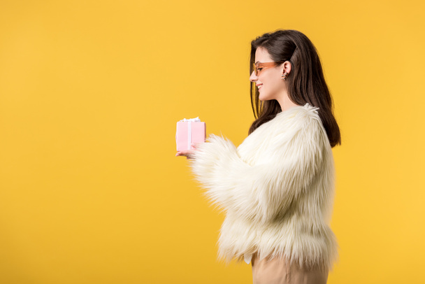 side view of happy party girl in faux fur jacket and sunglasses holding gift box isolated on yellow - Fotoğraf, Görsel