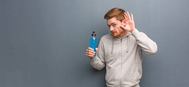 Young redhead fitness man try to listening a gossip. He is holding an energy drink. - Photo, Image