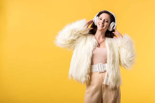 smiling girl in faux fur jacket listening music in headphones isolated on yellow - Φωτογραφία, εικόνα