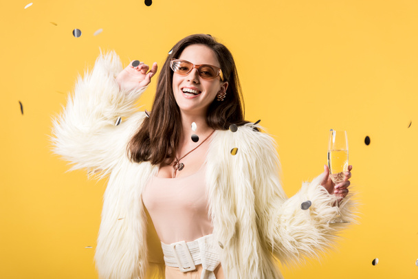 excited party girl in faux fur jacket and sunglasses standing under confetti with champagne isolated on yellow - Foto, imagen
