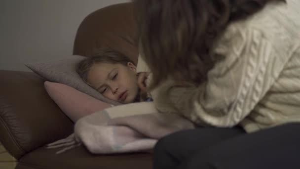 Молодая брюнетка в белом свитере измеряет температуру своей больной дочери дома. Маленькая симпатичная кавказка лежит под теплым одеялом с лихорадкой
. - Кадры, видео
