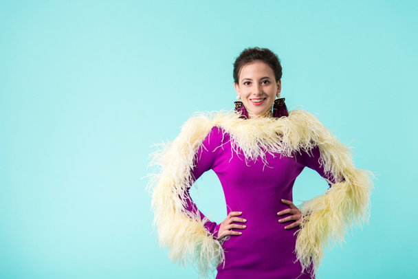 happy party girl in purple dress with feathers standing with hands on hips isolated on turquoise  - Photo, image