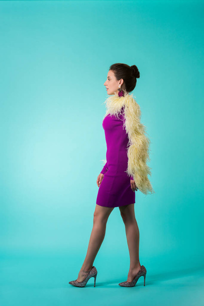 vista lateral de la chica de la fiesta en vestido púrpura con plumas caminando sobre fondo turquesa
 - Foto, Imagen