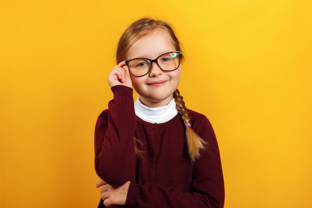 Clever little girl school girl corrects glasses. A child in a red sweater on a yellow background - Fotó, kép