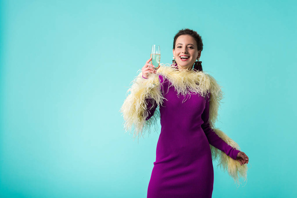 happy party girl in purple dress with feathers holding glass of champagne isolated on turquoise  - Φωτογραφία, εικόνα