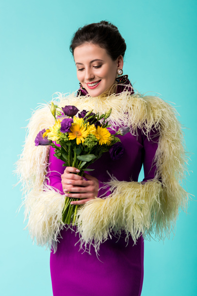 smiling party girl in purple dress with feathers holding bouquet of flowers isolated on turquoise  - Photo, image